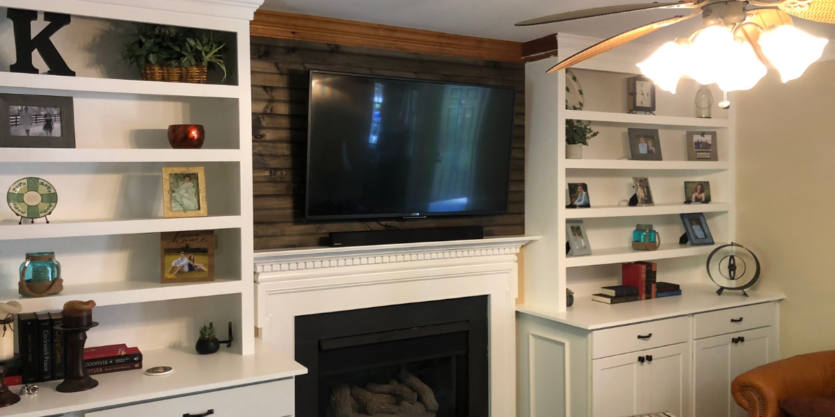 Living room with white built-in shelves on either side of a white fireplace surround, a mounted flat-screen TV above the fireplace, and various decorative items and framed photos on the shelves. A ceiling fan with lights is partially visible at the top right corner.