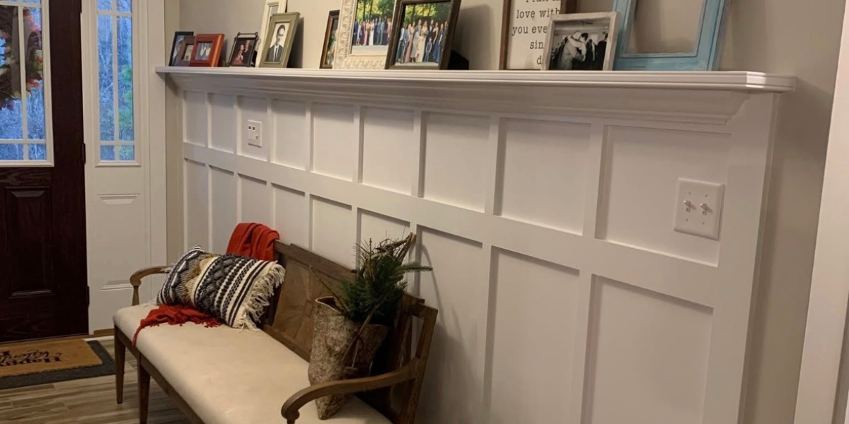 Hallway with a white wainscoting wall, decorated with family photographs on a ledge, a wooden bench with pillows and a red scarf, and a small potted plant. A door with glass panels is partially visible on the left.