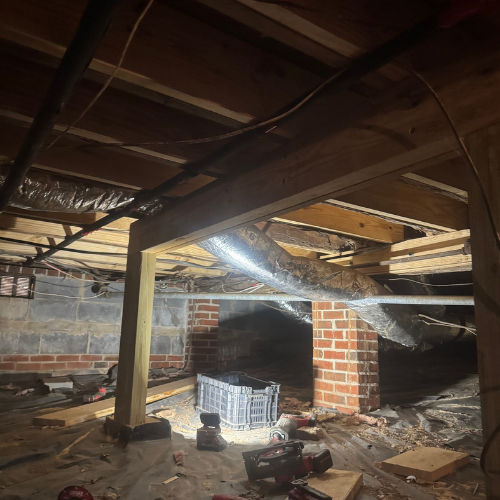 View of an unfinished basement or crawl space with exposed wooden beams, brick supports, and electrical wiring overhead. Construction tools and supplies are scattered on the floor, and a large duct runs across the space.