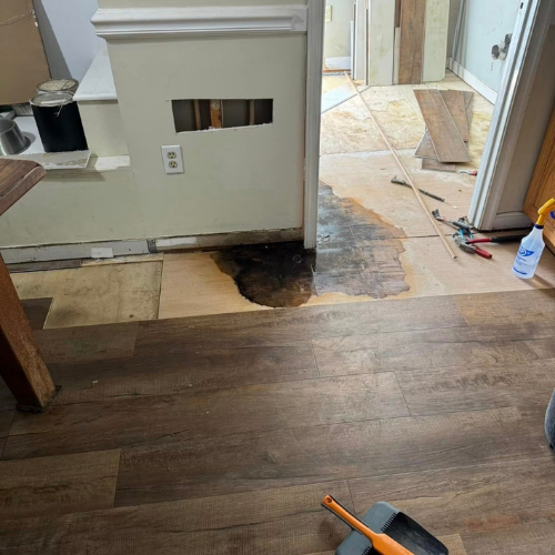 Room with partially installed wooden flooring, tools, and construction materials, showing a wet patch on the floor near the wall.