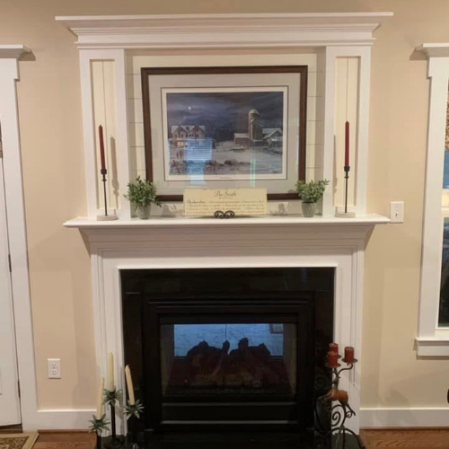 Decorative white fireplace mantel with framed landscape painting, two red candles, two potted plants, and a small paper sign, with a black fireplace insert below and a window to the right.