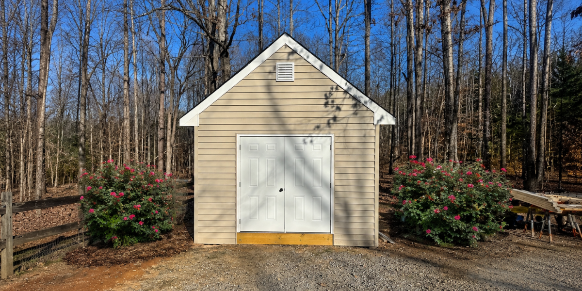 Small beige shed with white double doors, surrounded by pink flowering bushes, set against a wooded background under a clear blue sky.