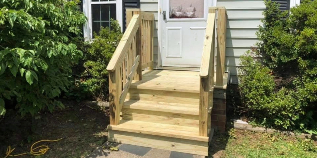 Newly built wooden ramp with handrails leading to a white door on the side of a house surrounded by green bushes.