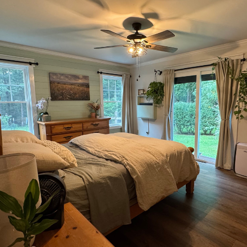 A bedroom with a bed covered in beige bedding, wooden furniture, and large windows with curtains showing a green yard outside.