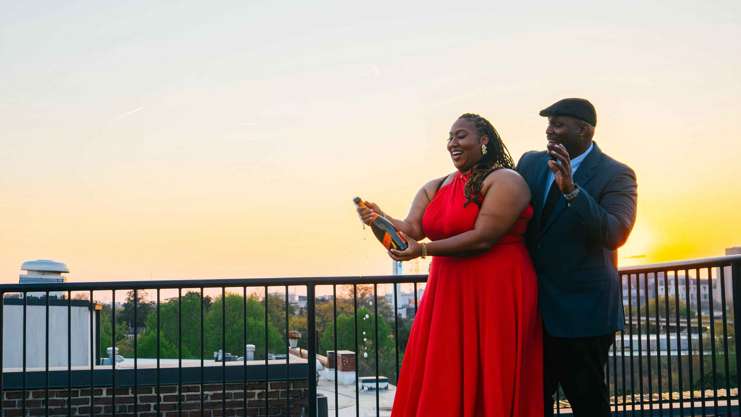 A woman in a red dress and a man in a suit and hat are standing on a rooftop balcony during sunset, smiling and holding a bottle of champagne.