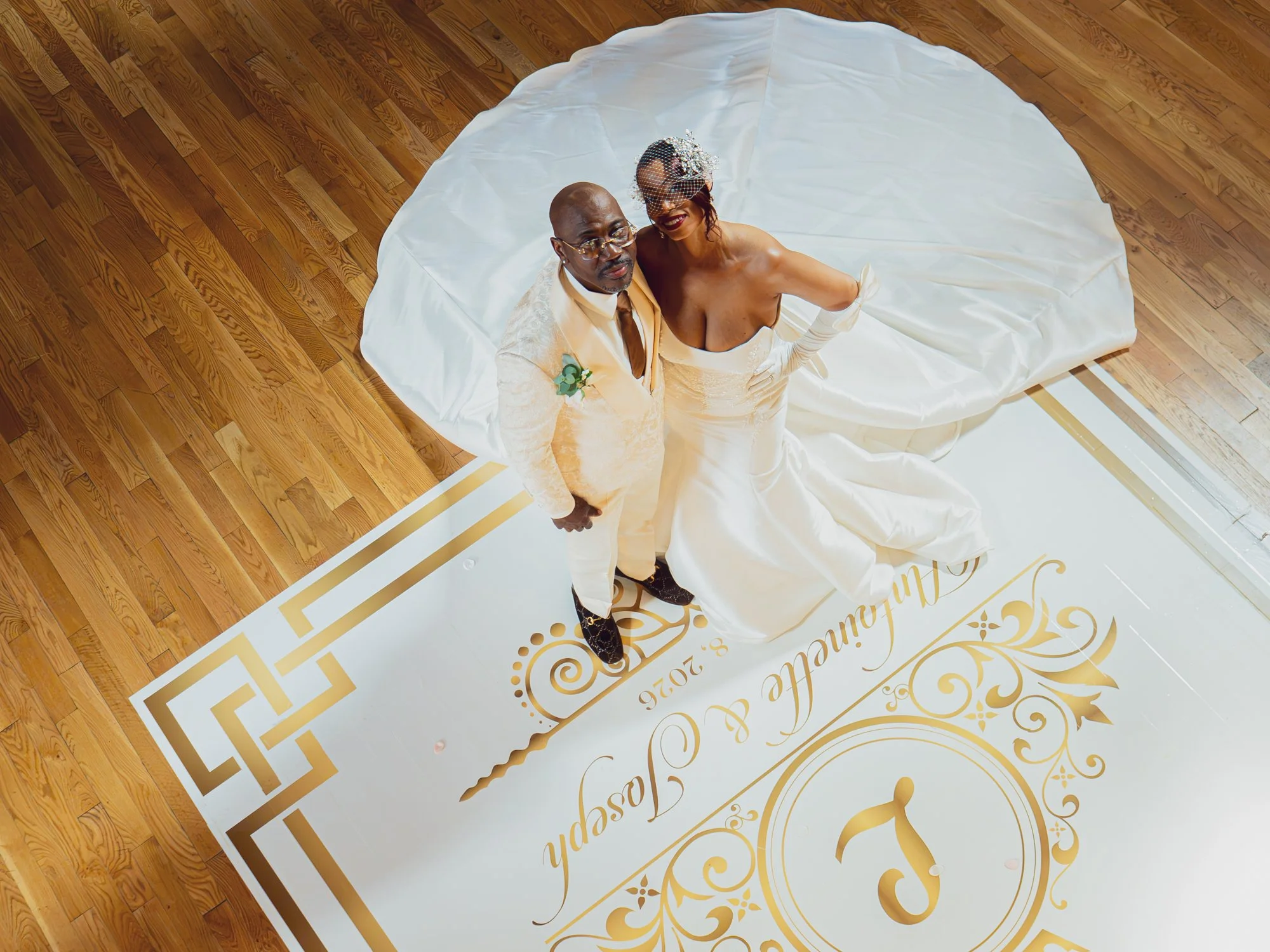 A newlywed couple standing on a decorated wedding backdrop, looking up at the camera, with the bride in a white gown and veil, and the groom in a beige suit, holding hands.