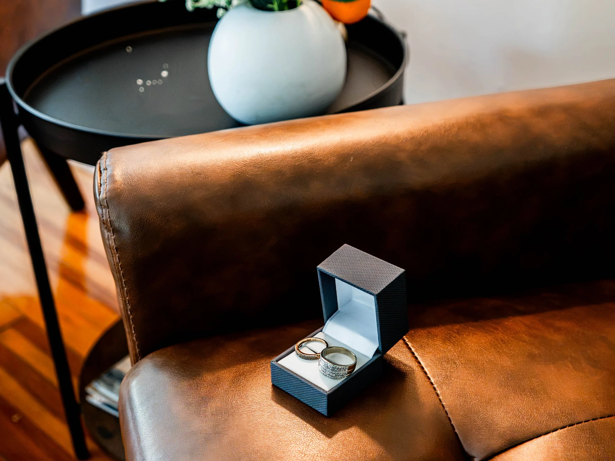A black jewelry box holding three rings, placed on a brown leather chair with a round back. In the background, a small potted plant sits on a black tray, and a pair of rings is on the chair's armrest.