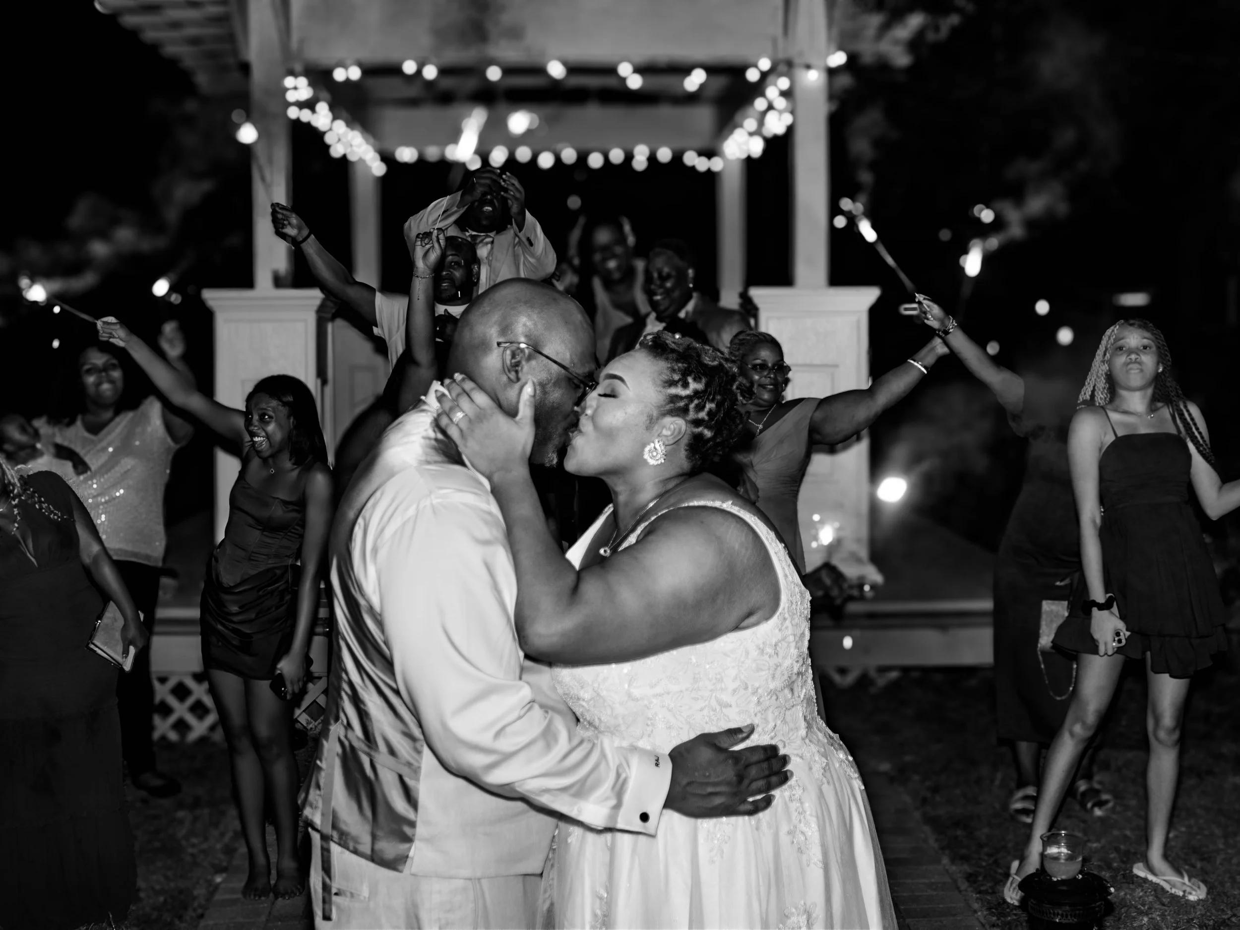 A black and white photo of a wedding reception outside at night. A bride and groom are kissing while surrounded by guests. String lights hang above, and guests are dancing and celebrating around the couple.
