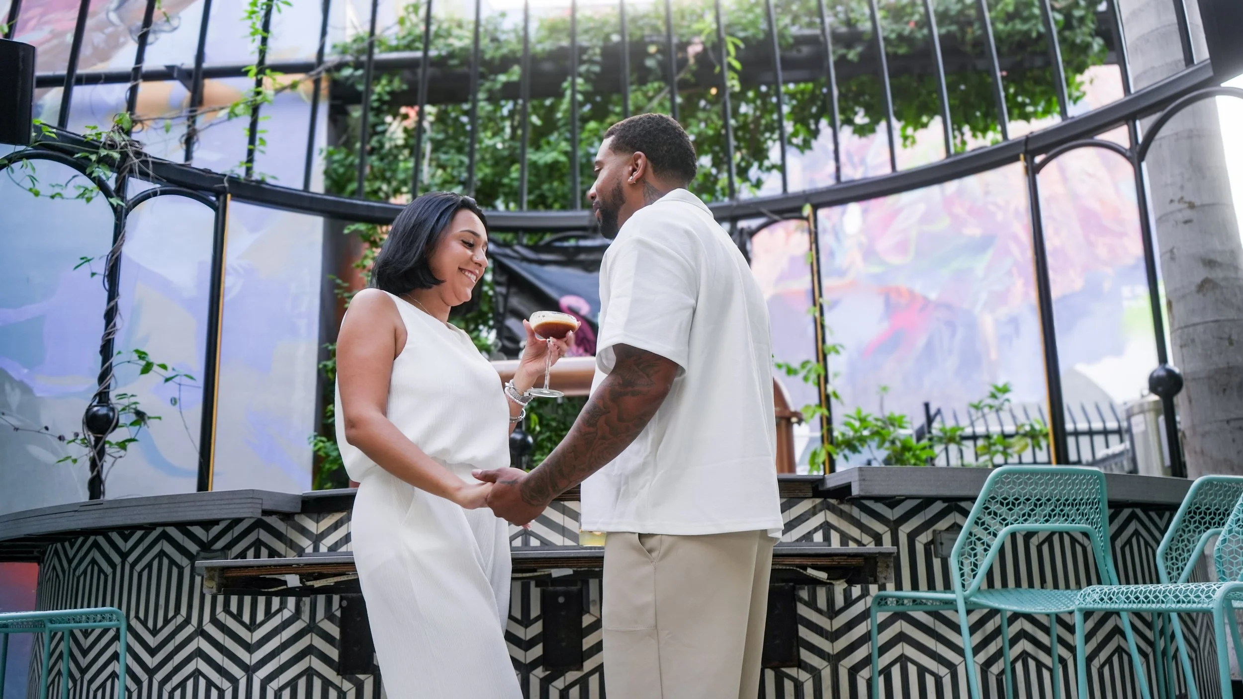 A couple holding hands, standing close together outdoors, exchanging a drink, smiling, with greenery and modern outdoor furniture in the background.