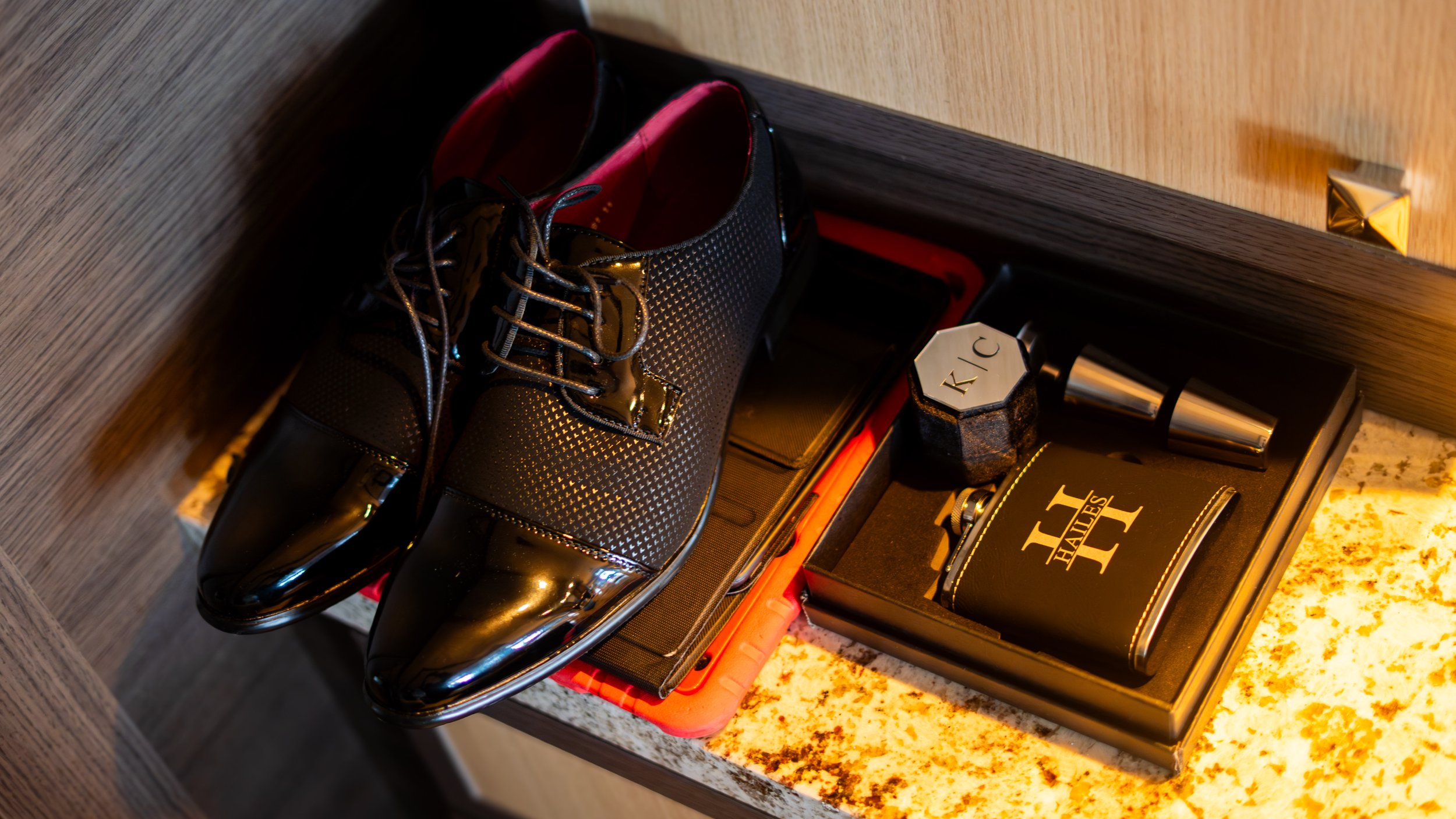 A pair of glossy black dress shoes with dotted pattern and laces, placed on a pink tray inside a drawer containing perfume bottles, a watch, and a black box labeled 'HIT'.