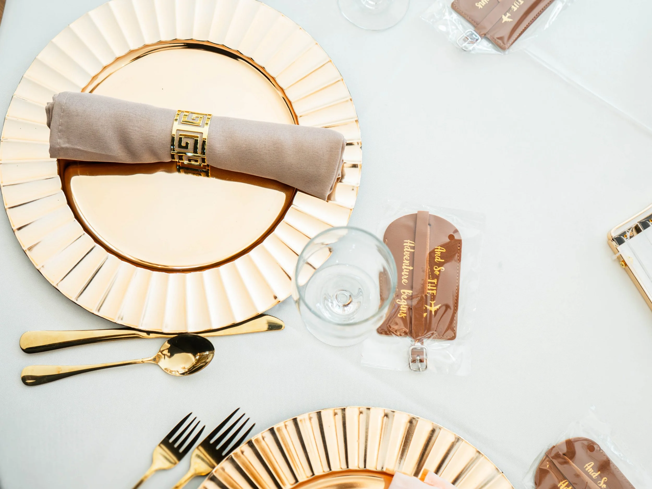 Elegant table setting with gold and beige plates, gold utensils, a beige napkin with a gold decorative ring, clear glasses, and brown goody bags with gold writing.