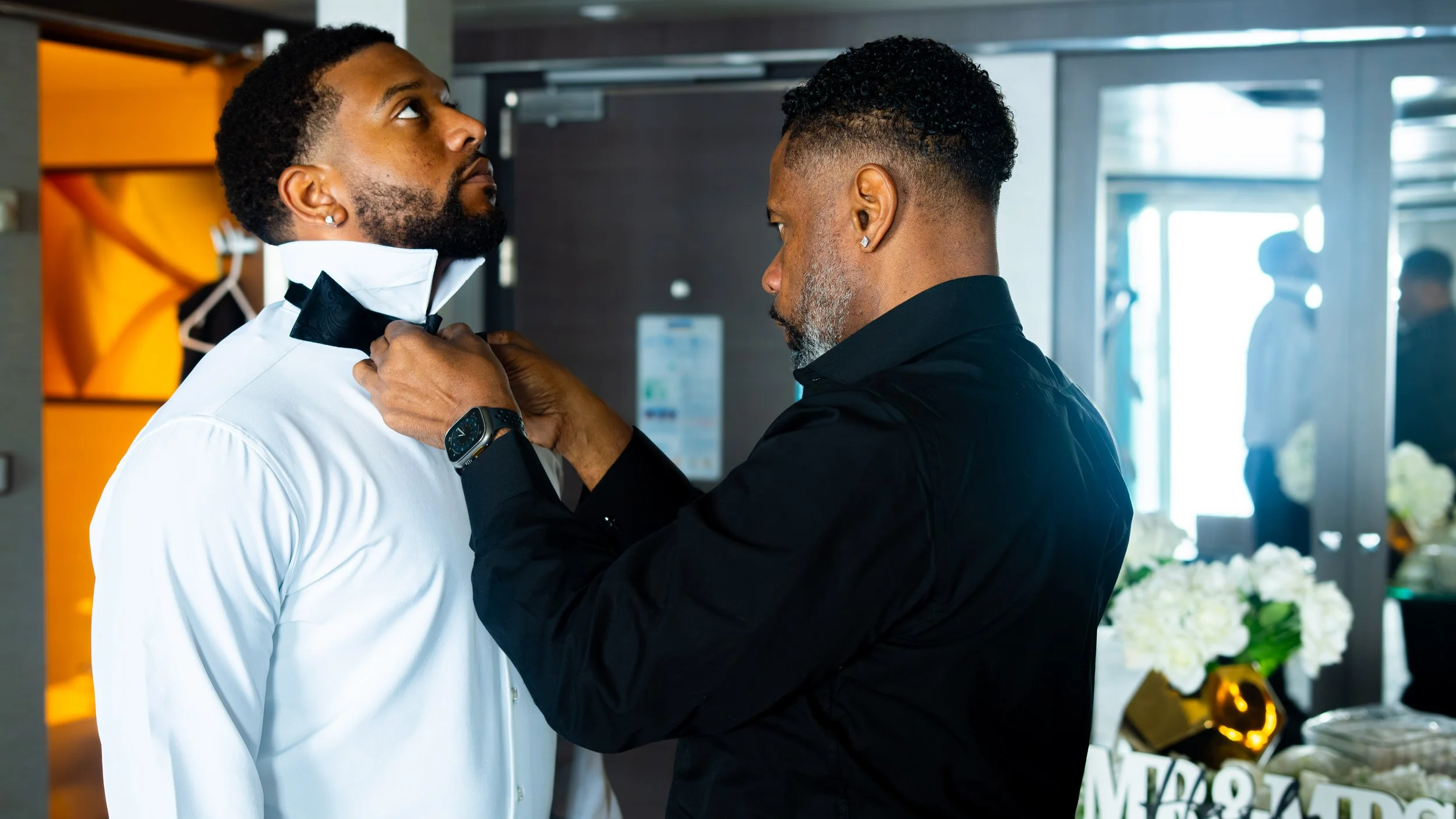 A man in a white tuxedo is having his bow tie adjusted by another man dressed in black. The setting appears to be indoors, possibly at a formal event.