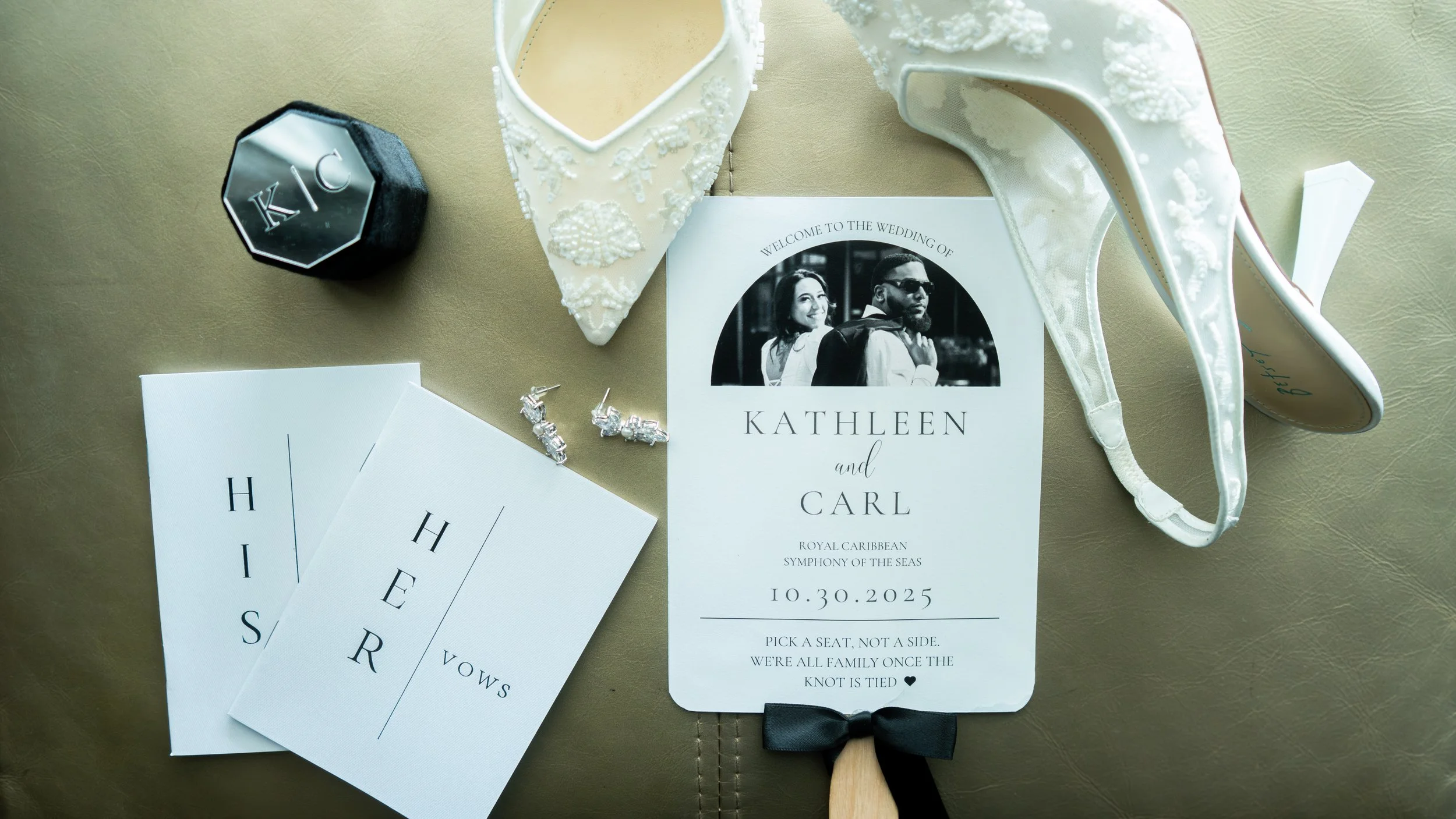 Wedding invitation surrounded by wedding accessories including a tuxedo, a pair of wedding shoes, diamond earrings, a black hexagonal box with initials K and C, and reception cards, on a beige background.