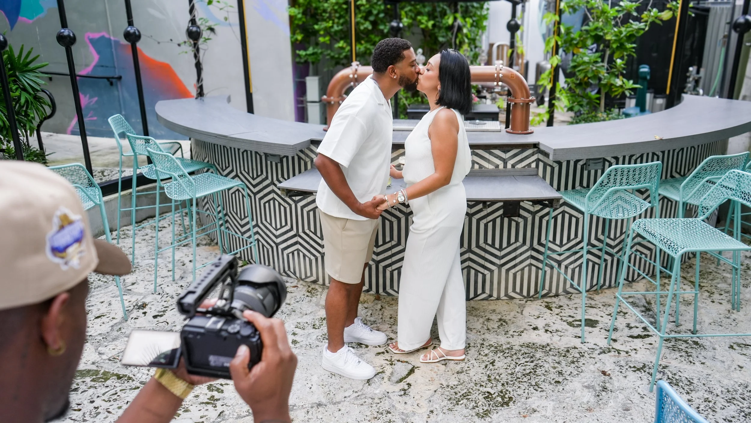 A couple is kissing in an outdoor setting surrounded by modern chairs and a bar with a geometric black and white pattern, with greenery in the background. A woman is taking a photo or video of the couple with a camera.