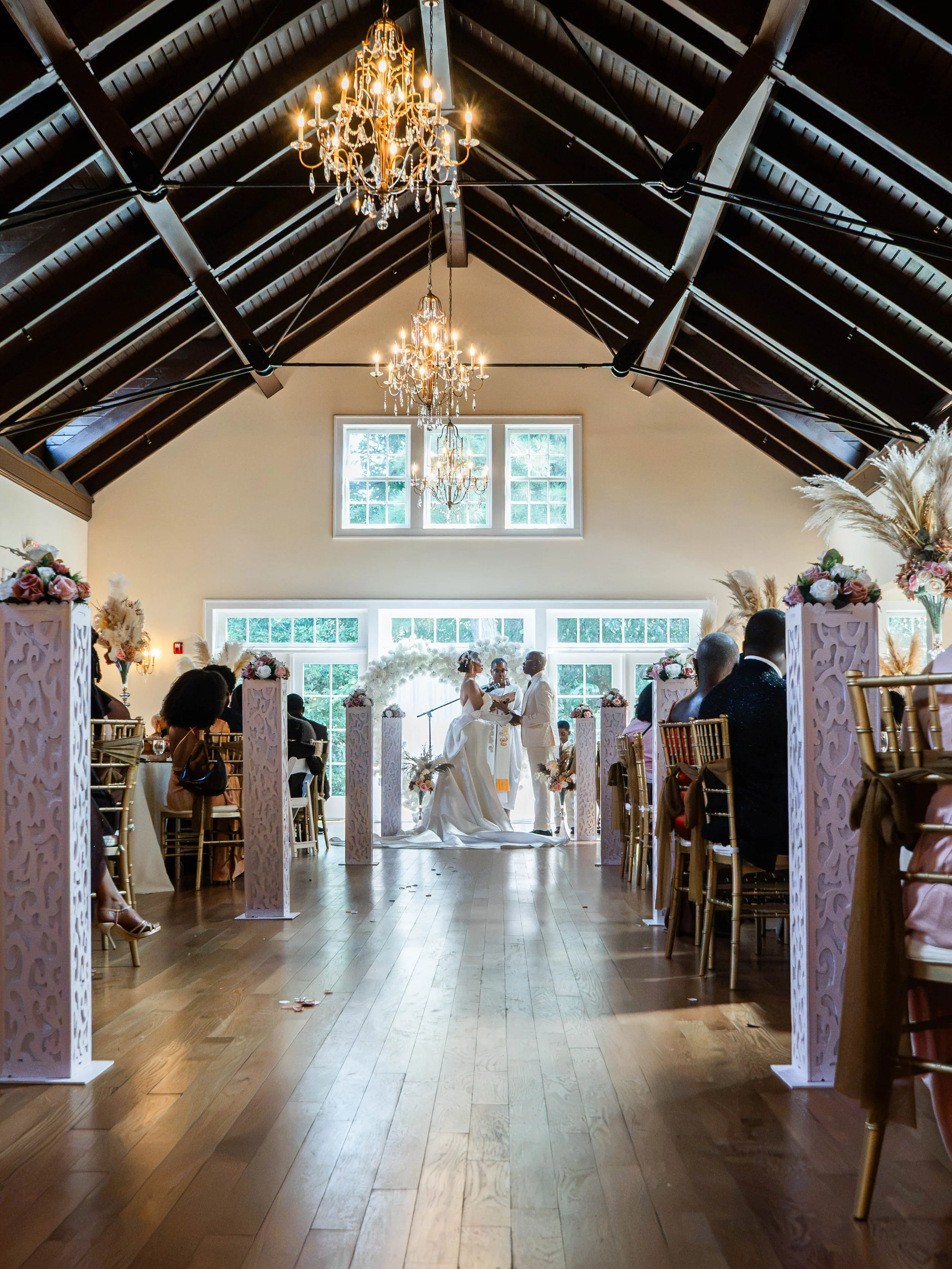 Interior of a wedding venue with high vaulted ceiling, wooden beams, chandeliers, and large windows in the background. A couple is standing at the altar with guests seated on chairs on either side, and floral decorations are visible.