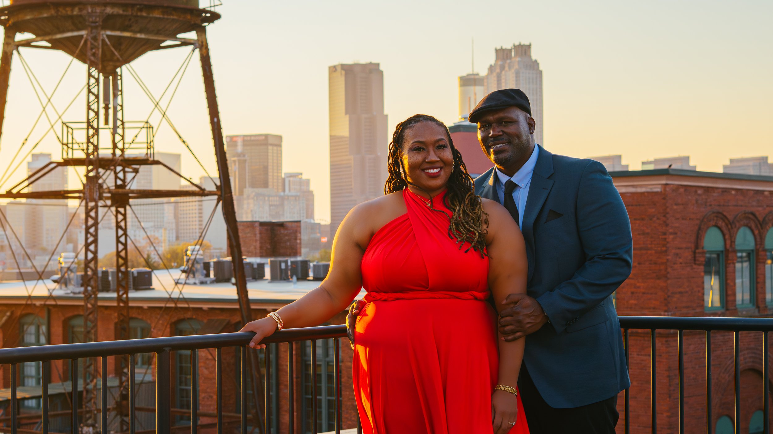 A smiling woman in a red evening gown and a man in a dark suit with a hat standing close together on a rooftop with city skyscrapers in the background during sunset.