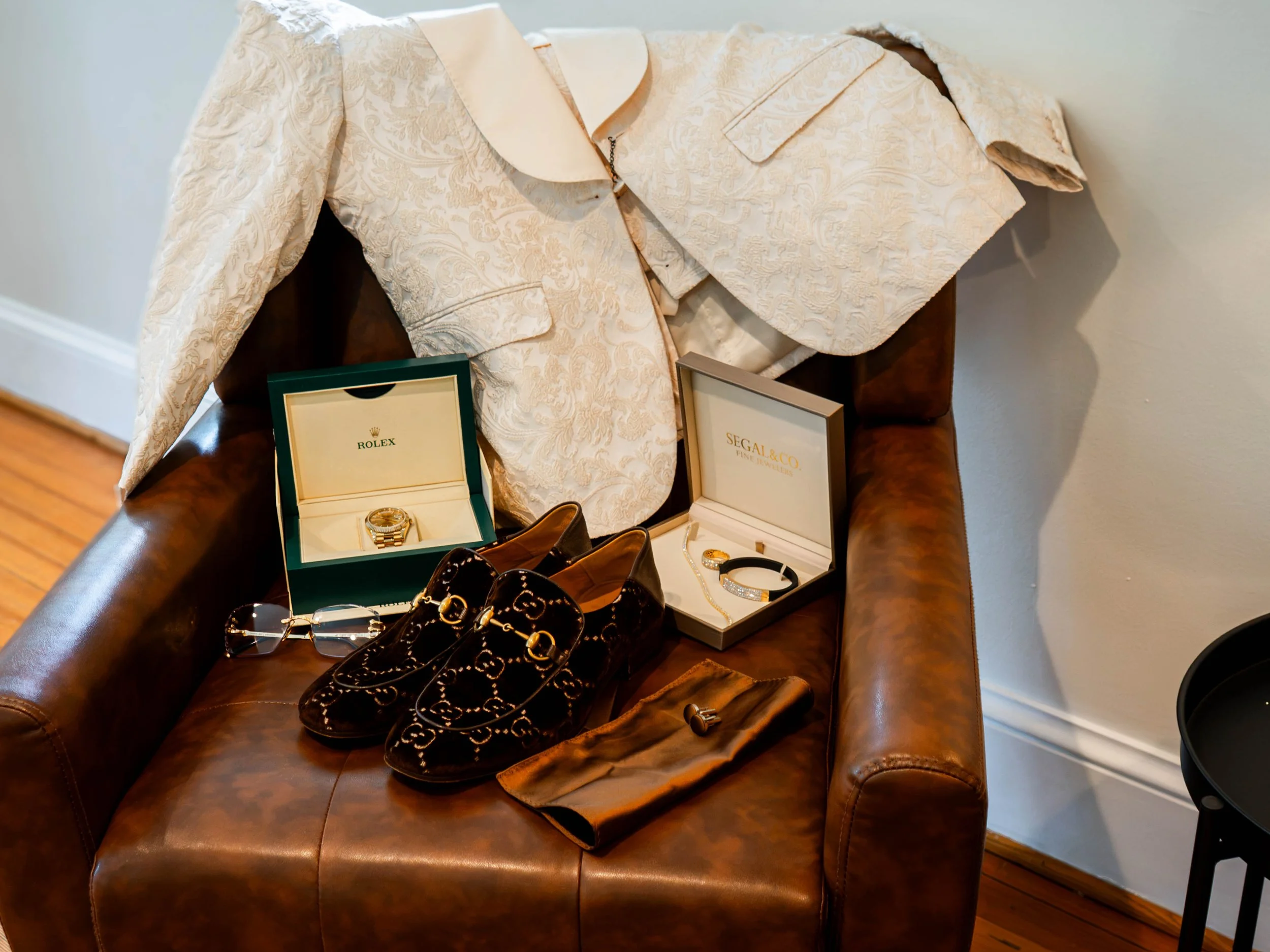 A brown leather couch with several pairs of shoes, jewelry, a watch, and boxed jewelry displayed on it, along with a tan jacket with a floral pattern hanging over the back of the couch.