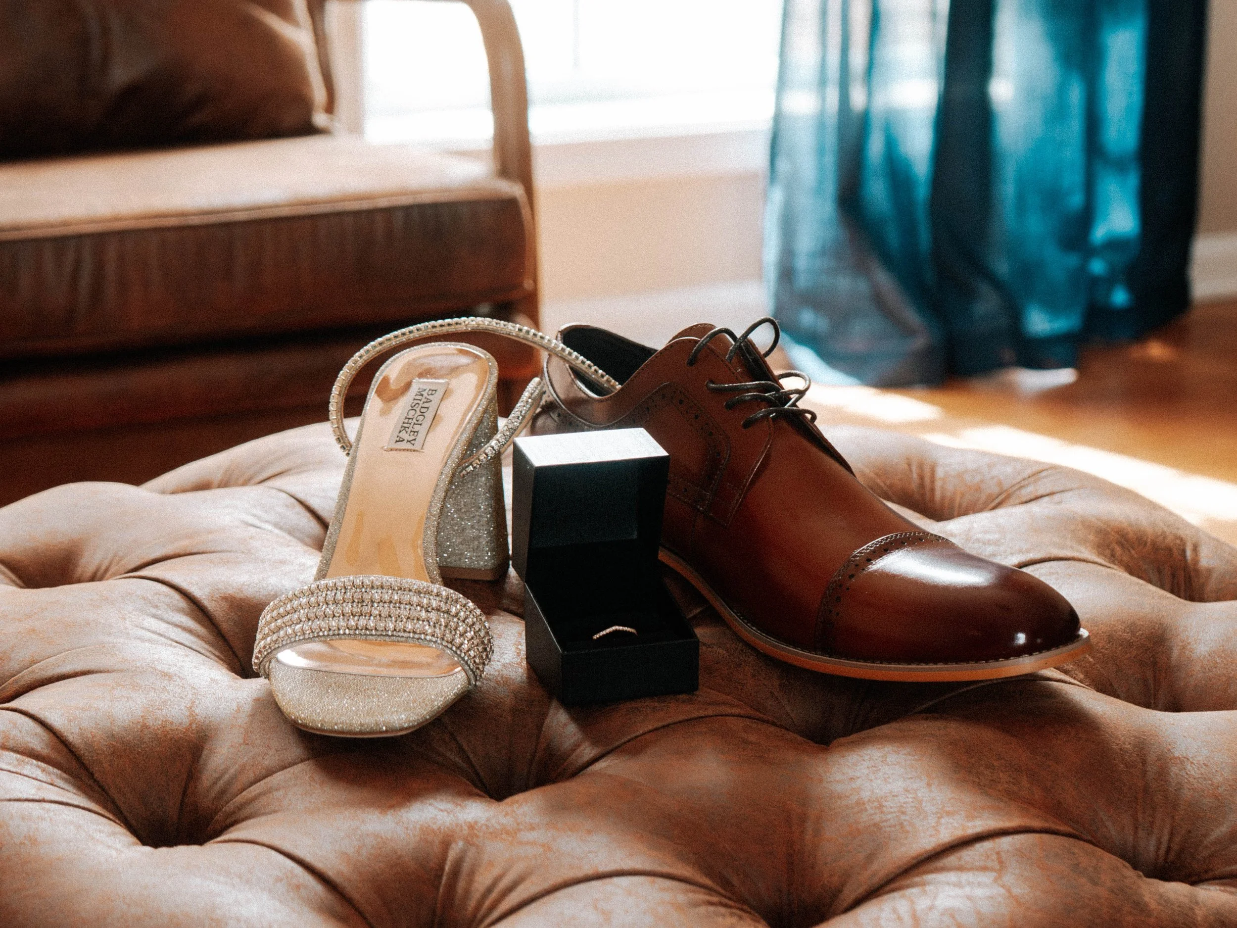 A pair of sparkly, gold-colored high heel shoes, a men's brown dress shoe, and a black box are placed on a tufted, blush-colored velvet surface. A bracelet and a box of jewelry are also visible.