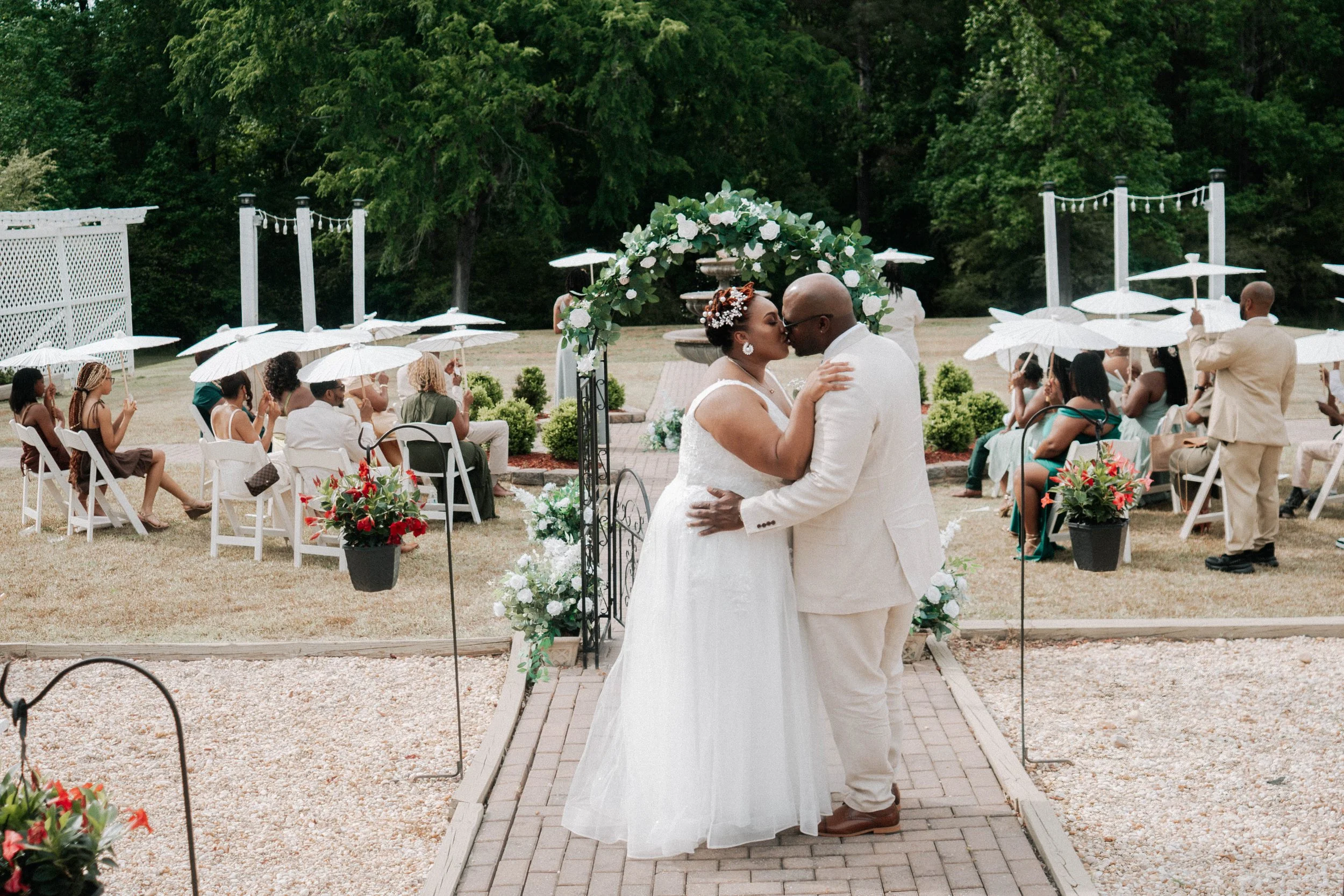 A couple sharing a kiss at their outdoor wedding ceremony, with guests seated nearby holding white umbrellas, and a decorative floral arch behind them.