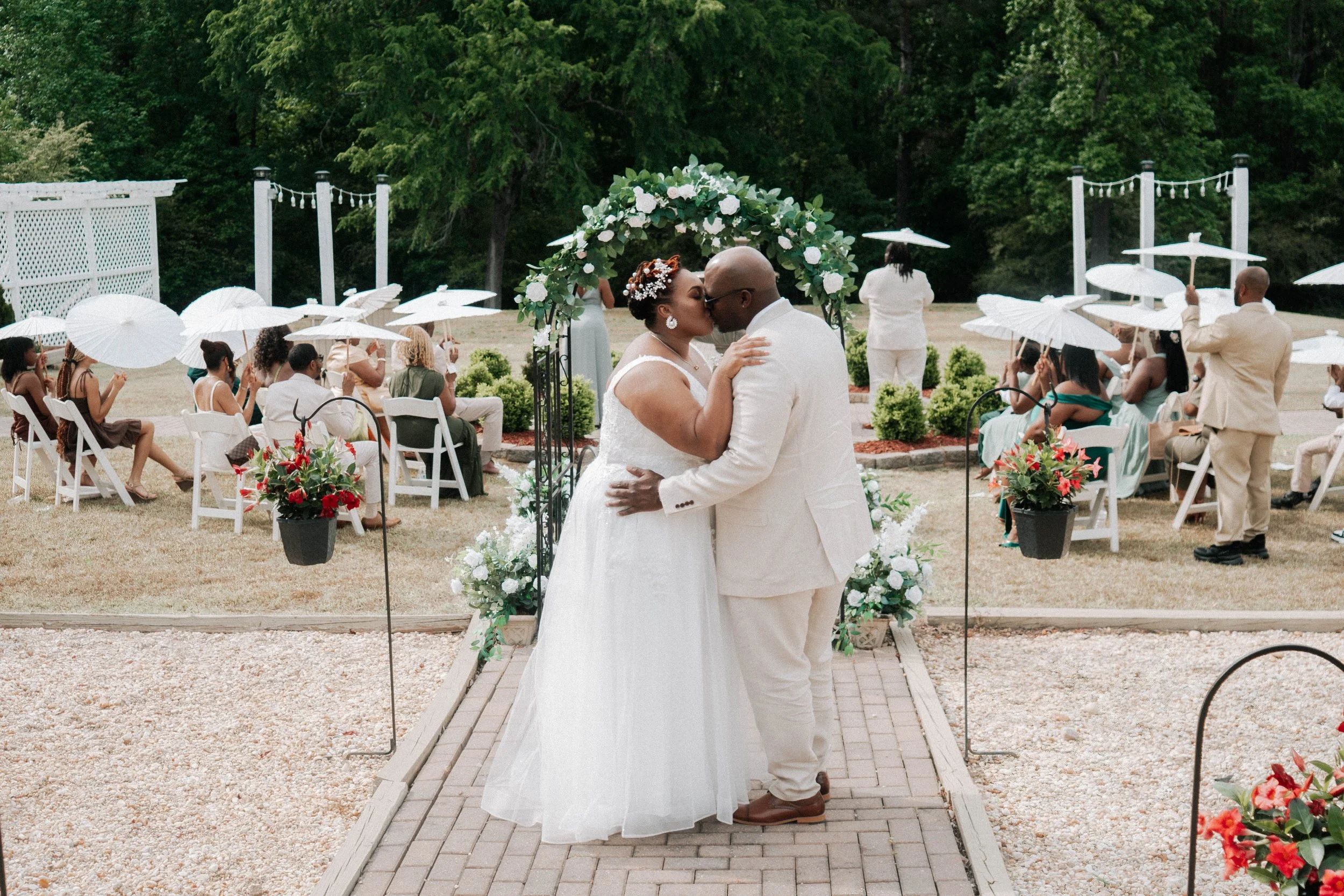 A newlywed couple sharing a kiss during their outdoor wedding ceremony, surrounded by guests holding white umbrellas, with a floral arch and lush trees in the background.