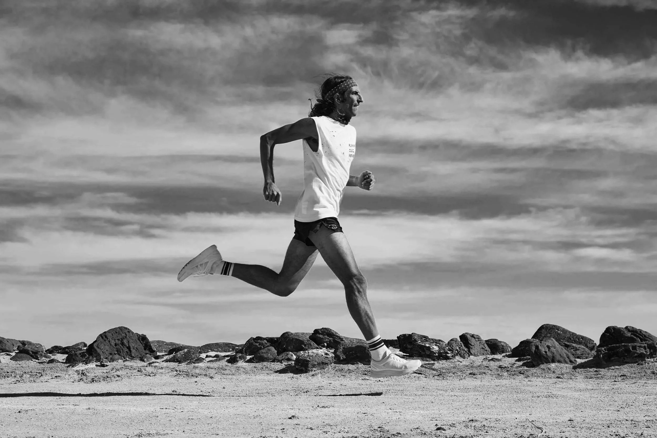 A person running on a rocky beach with a cloudy sky in the background, wearing a tank top, shorts, and running shoes.