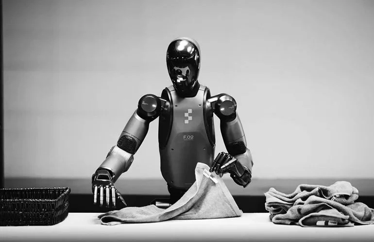 A humanoid robot with a black head and metallic body folding laundry on a table.