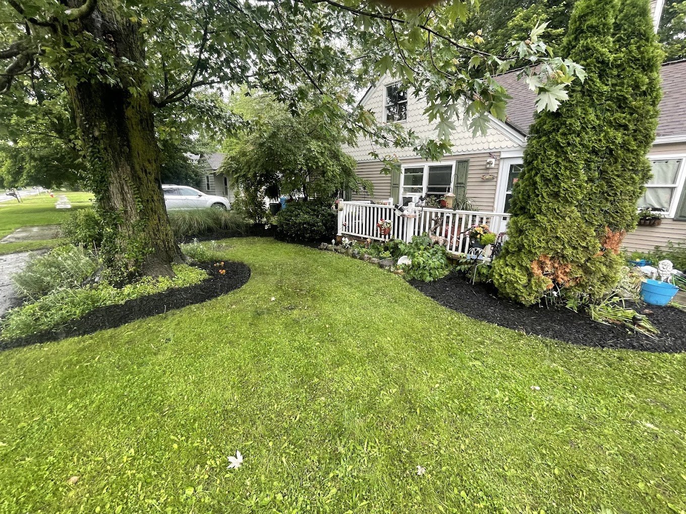 A well-maintained front yard with a large tree, lush green grass, and flower beds with decorated plants and garden ornaments, in front of a house with a small porch.