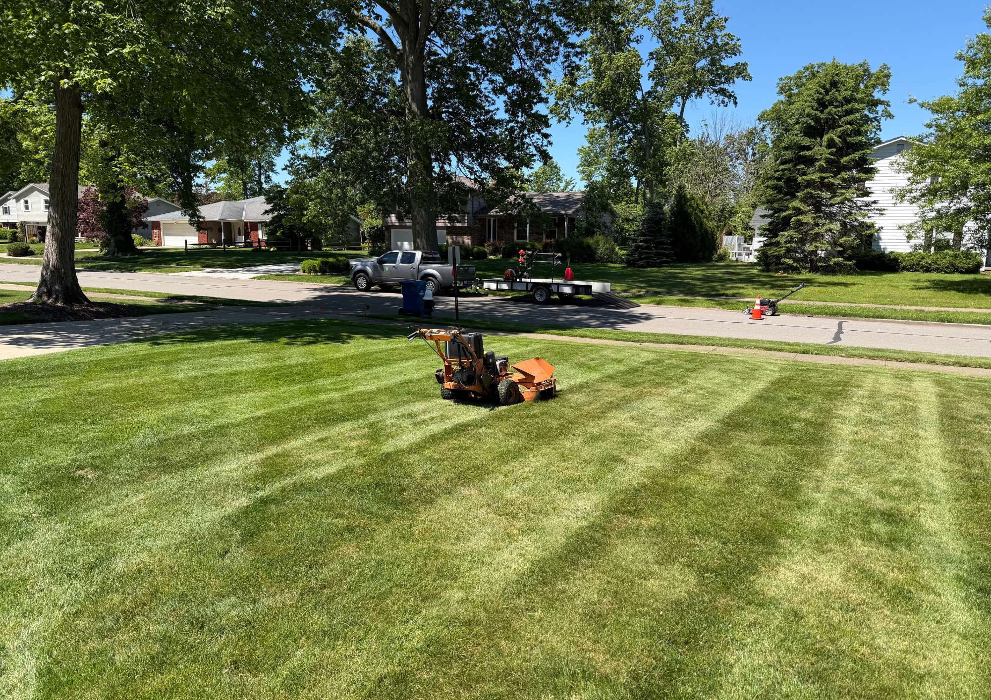 A suburban residential street with trees, houses, a pickup truck, a trailer, a lawn mower, and a traffic cone on a sunny day.