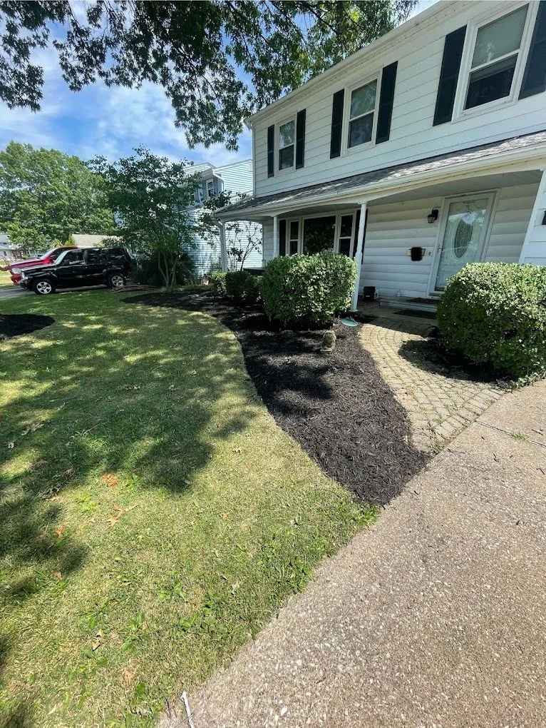 Front yard of a house with a newly landscaped garden bed with mulch, green bushes, a brick pathway, a white house, and parked cars in the background.