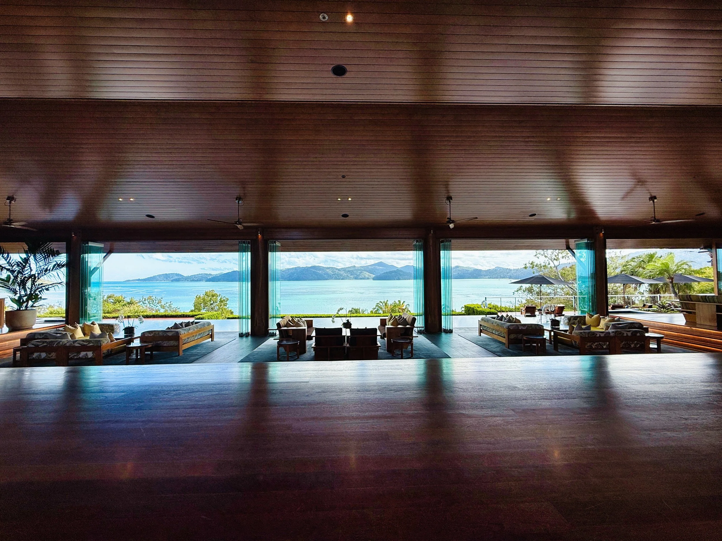 View of a spacious indoor lounge area with wooden ceiling, large open windows, and a scenic ocean and mountain view outside, featuring seating arrangements and ceiling fans.