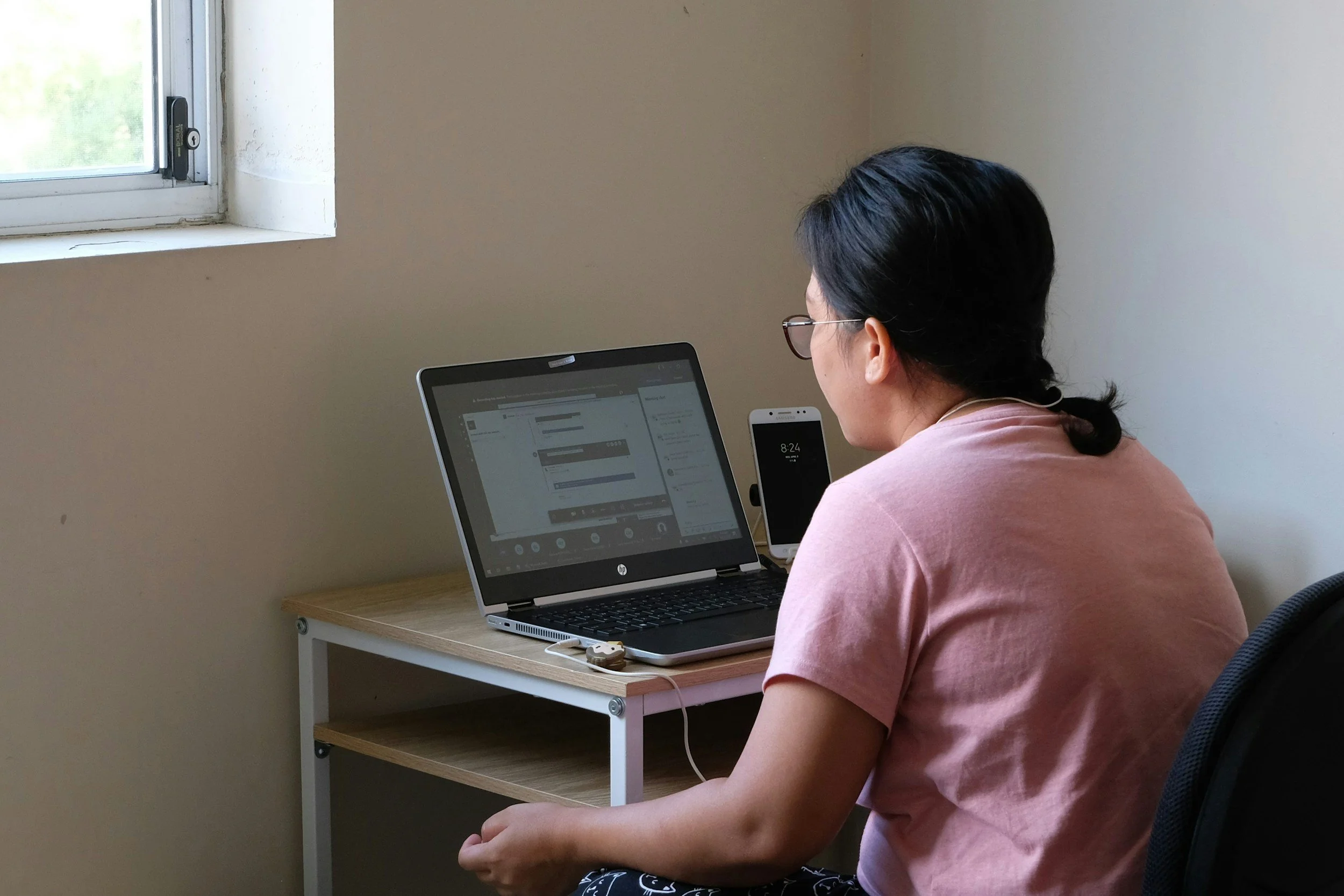 A woman with short dark hair and glasses is sitting at a desk using a laptop. The laptop is showing a video conference or an online meeting. There is a smartphone placed vertically on the desk next to the laptop, displaying the time 8:24. The desk is small with a lower shelf, and the wall behind is plain. There is a window on the left side letting in natural light.