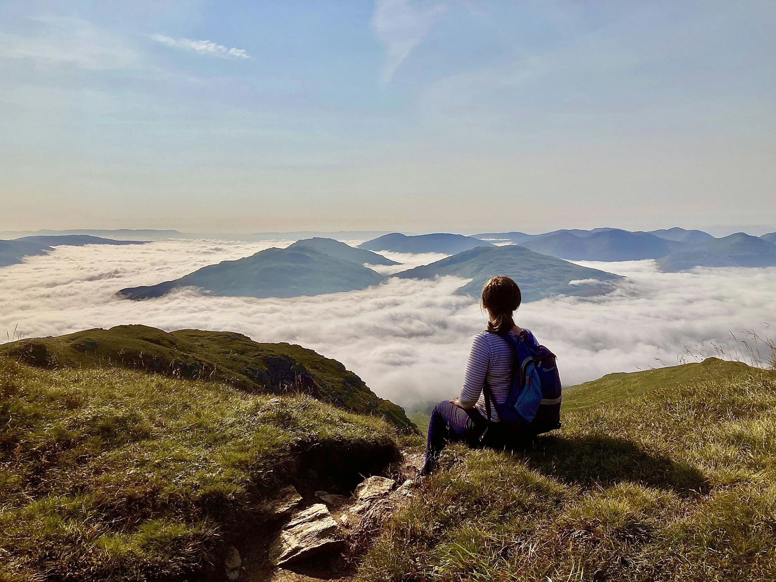 A person with a backpack sitting on the grassy ground on a mountain, overlooking clouds and mountains during daytime.