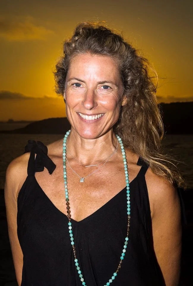 A smiling woman with curly hair standing outdoors during sunset, wearing a black sleeveless top with tied shoulder straps and layered necklaces.