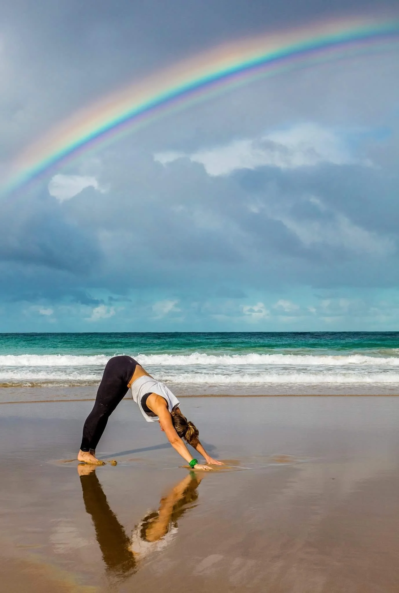A woman practicing yoga on the beach, performing a downward dog pose with the ocean and a rainbow in the background.
