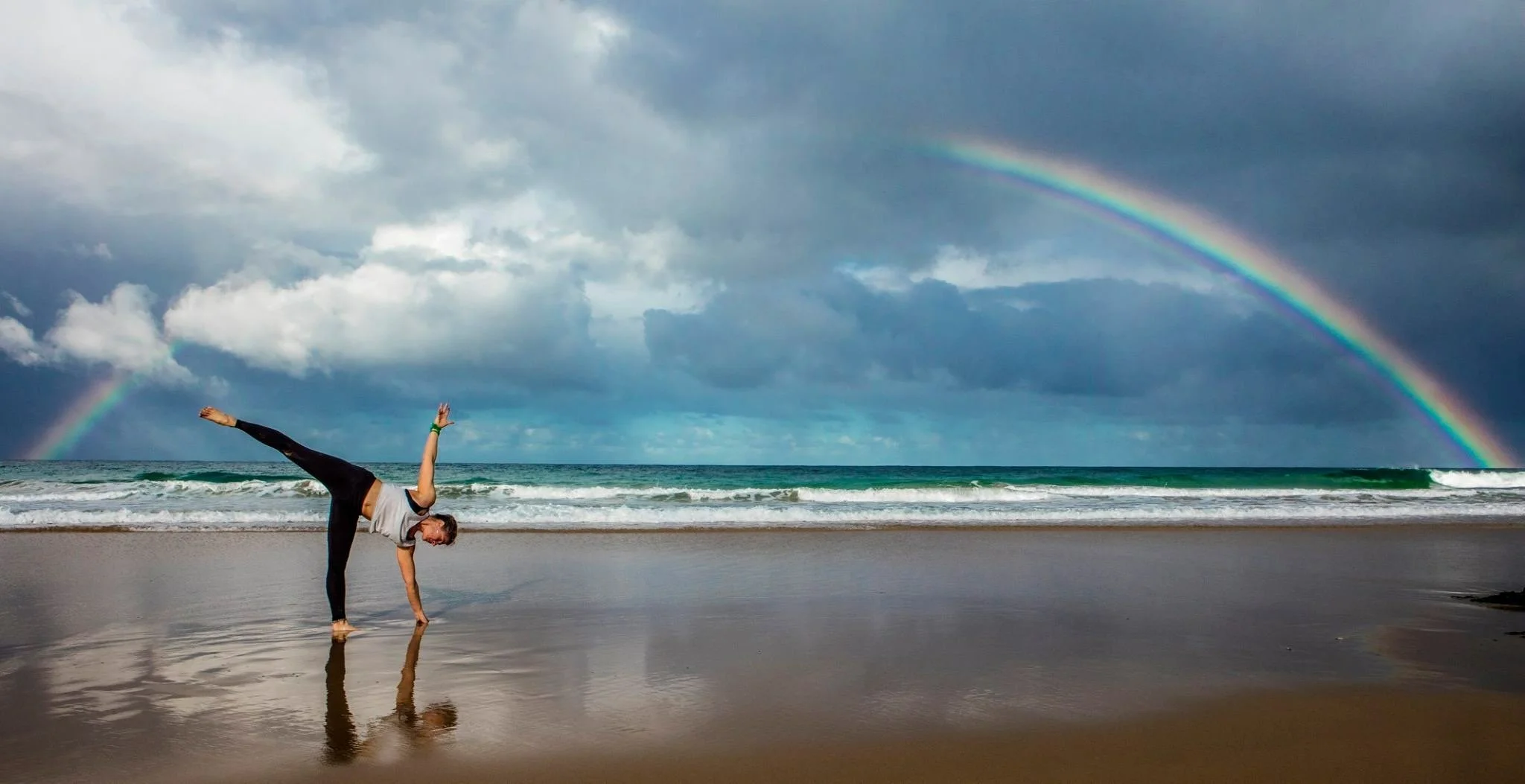 A person doing a handstand on a beach with a rainbow in the cloudy sky above ocean waves.