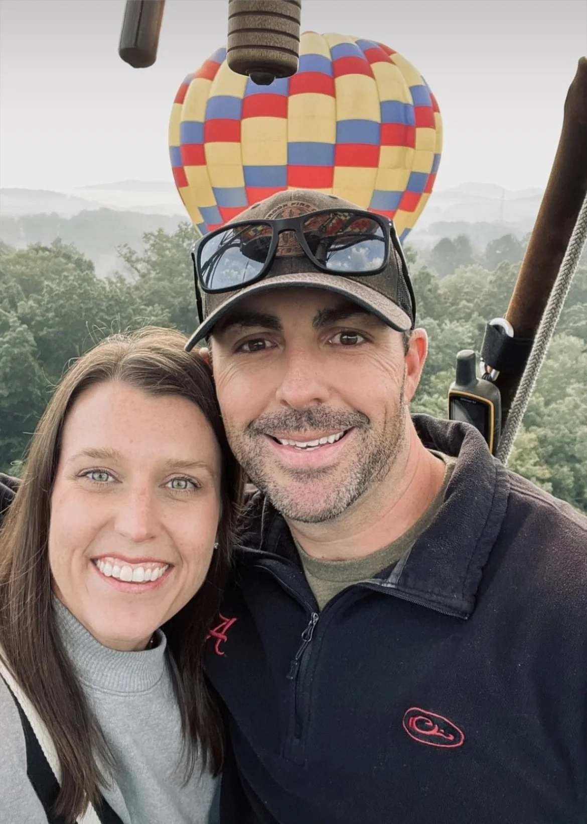 A couple taking a selfie in a hot air balloon, with the colorful balloon visible in the background and lush green trees below.