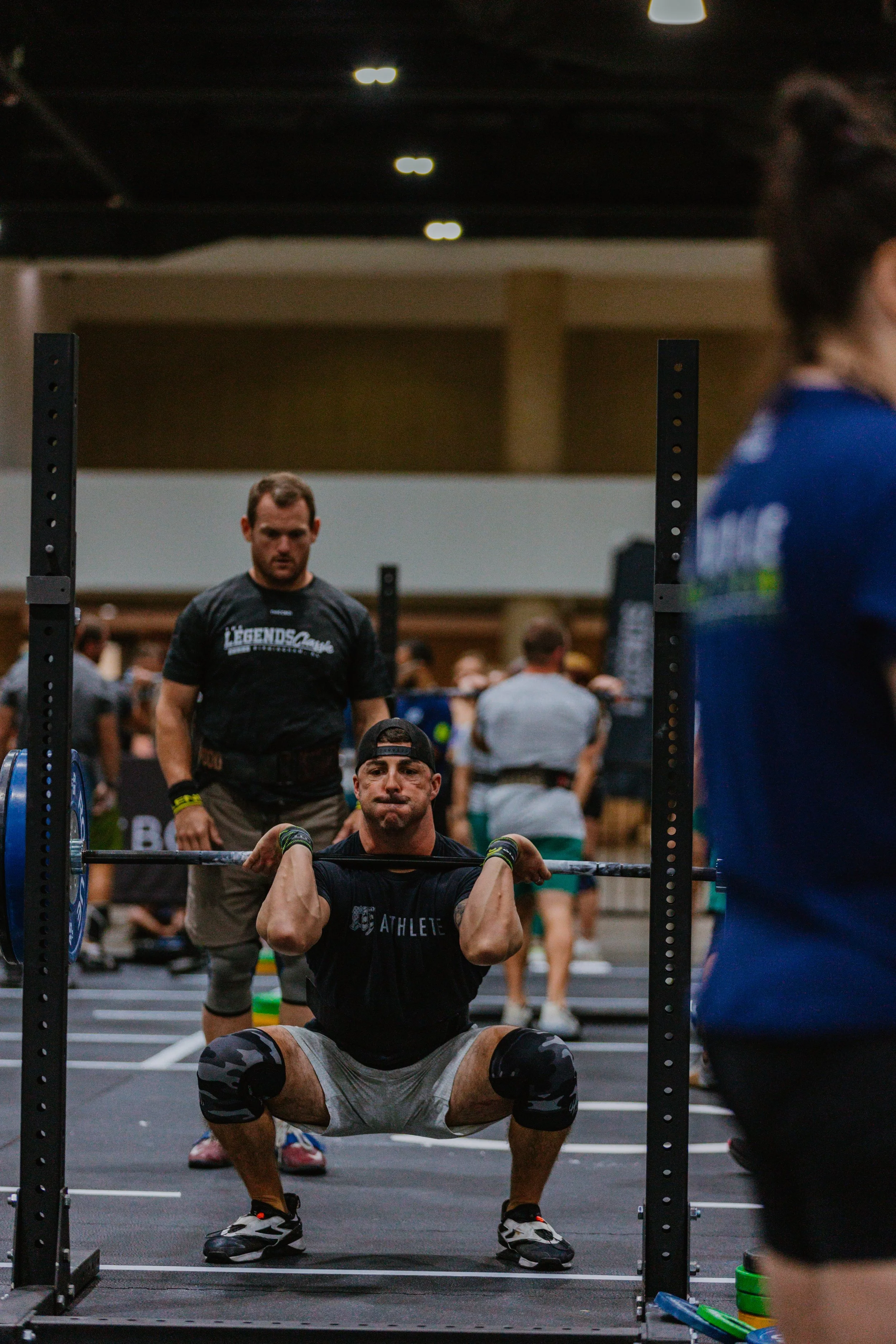 Man in black shirt doing squat exercise with barbell, surrounded by people in a gym, in a crowded fitness event.