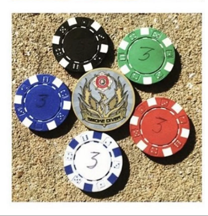 Six poker chips in black, green, blue, white, red, and gray with a central medal featuring a phoenix and the words "Game Over" on a sandy surface.