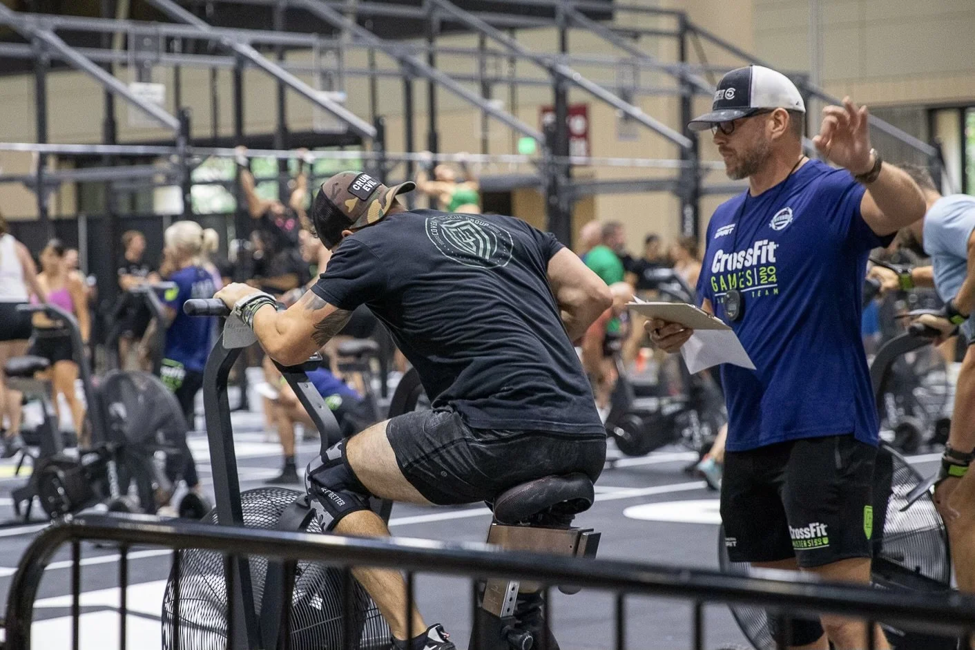 A man in black athletic gear riding a stationary bike at a CrossFit event, with a coach or trainer standing nearby taking notes, in a busy gym environment.