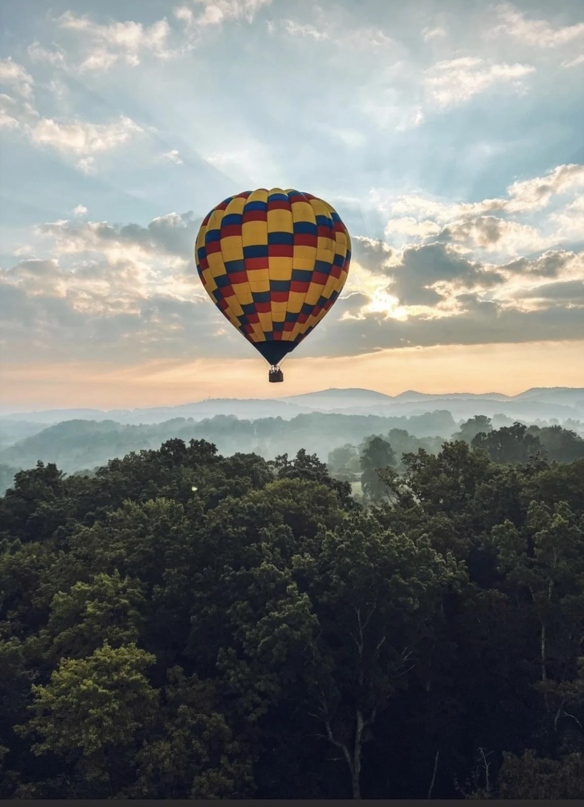 Colorful hot air balloon floating above a forested landscape at sunrise or sunset with a partly cloudy sky.