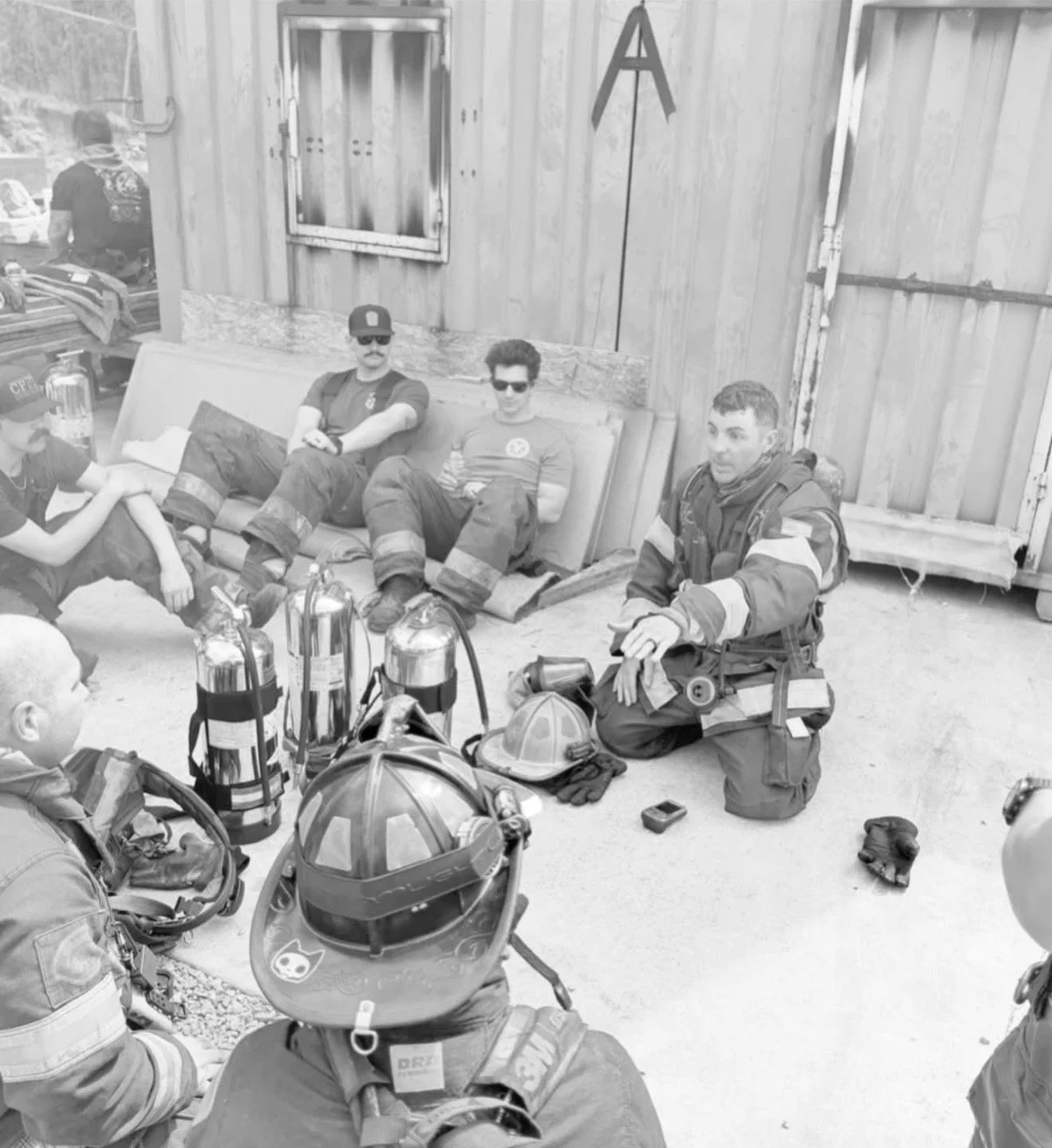 Firefighters and emergency personnel resting and having a conversation in a residential setting, with fire helmets and equipment visible.