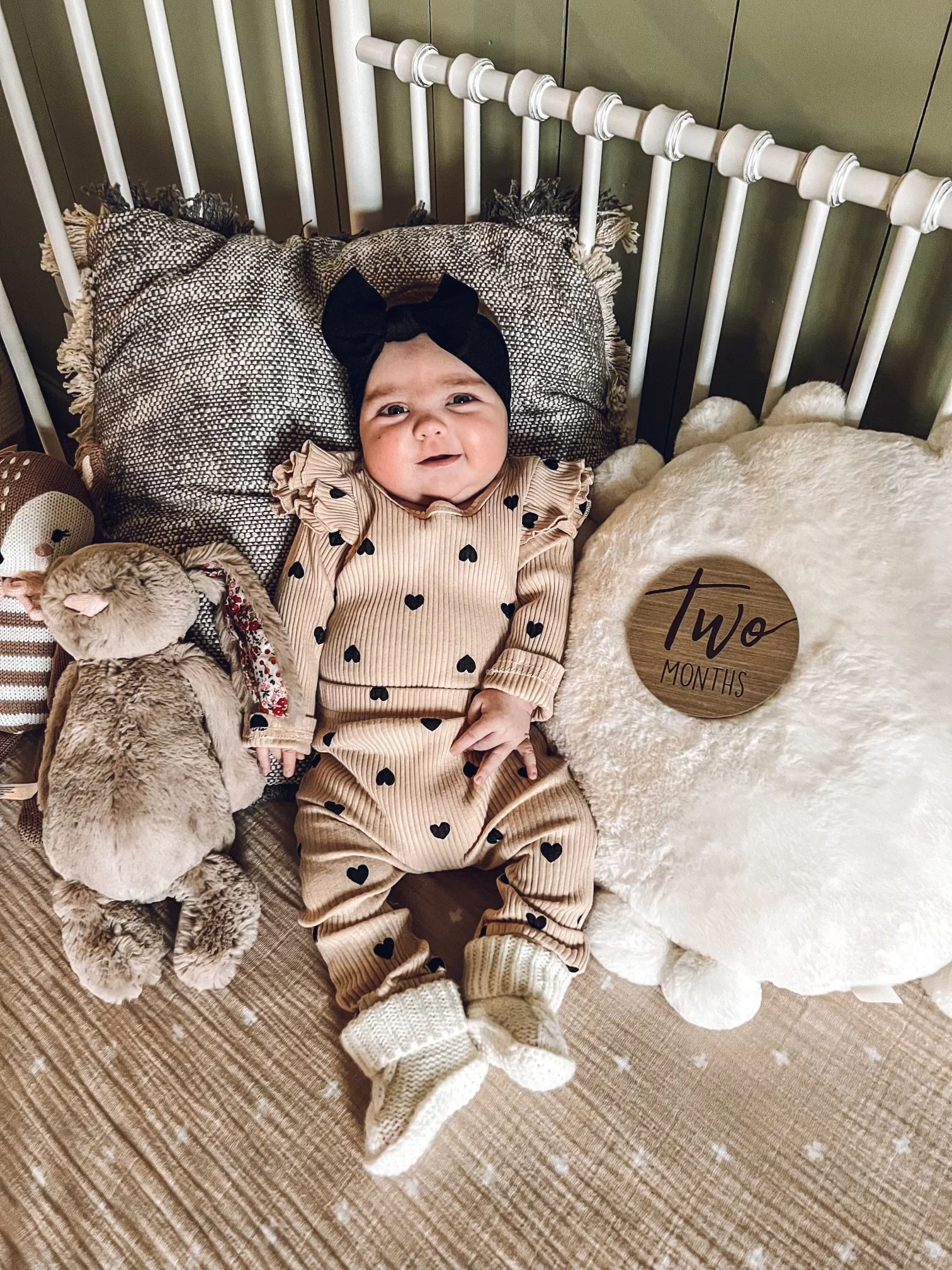 A smiling baby girl lying in bed with pillows, stuffed animals, and a sign that reads 'Two Months'.