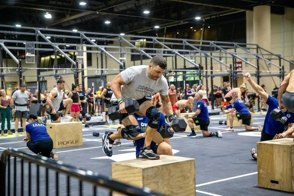 A man jumping onto a wooden box holding dumbbells in a busy gym during a CrossFit competition.