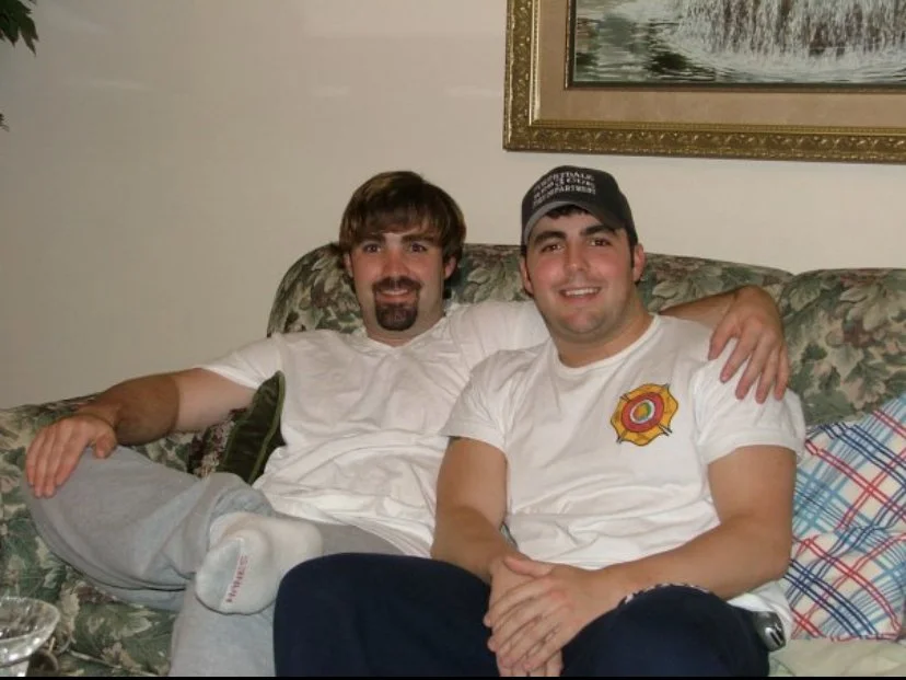 Two men sitting on a floral patterned couch, smiling, with one man’s arm around the other. There is a framed painting on the wall behind them.