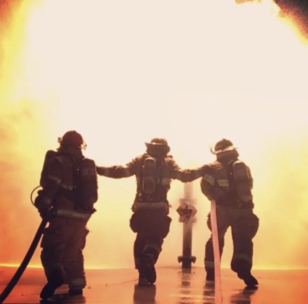 Three firefighters in protective gear stand in front of a large fire, holding hands, with the flames and smoke in the background.