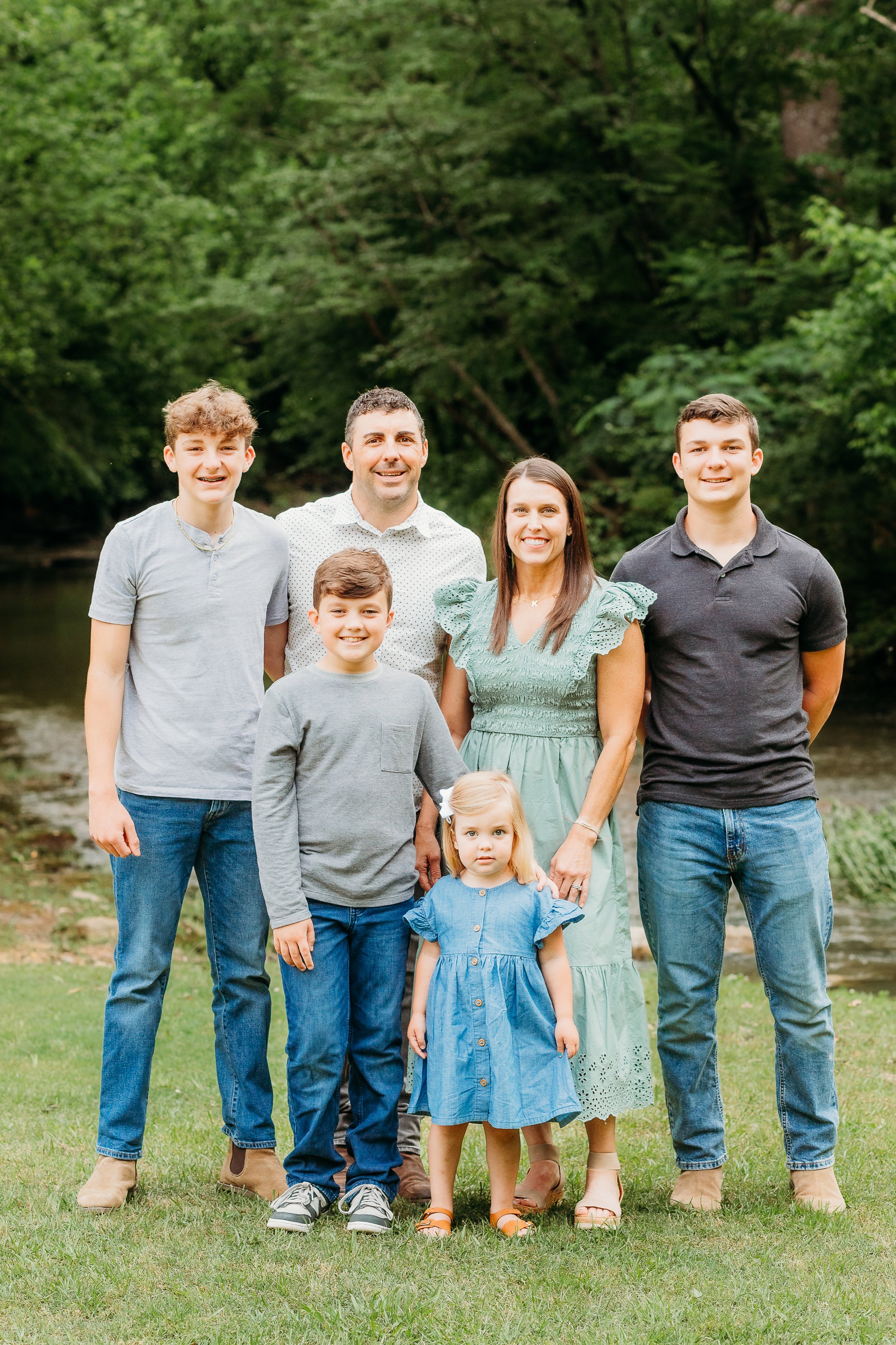 Family of eight people outdoors in a lush green park, standing on grass with trees in the background, smiling at the camera.