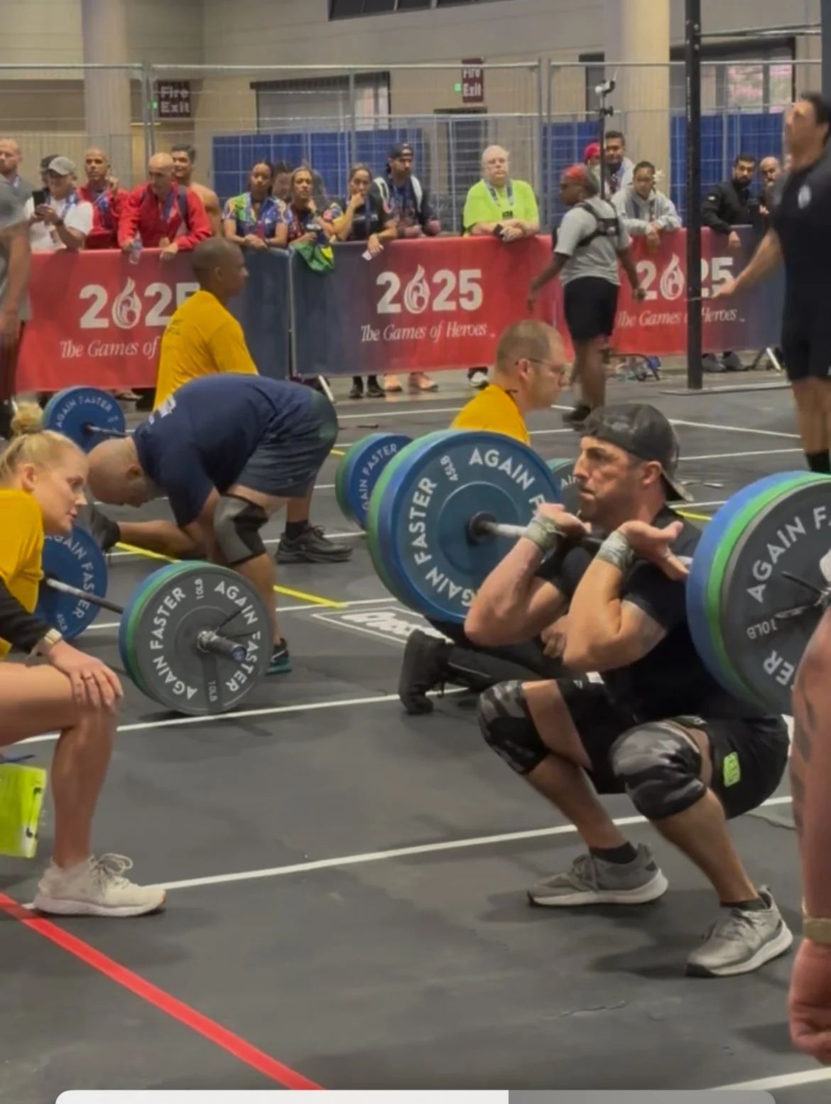 A man squats with a loaded barbell during a weightlifting competition at the 2023 Games of Heroes event, with spectators watching behind a barrier.