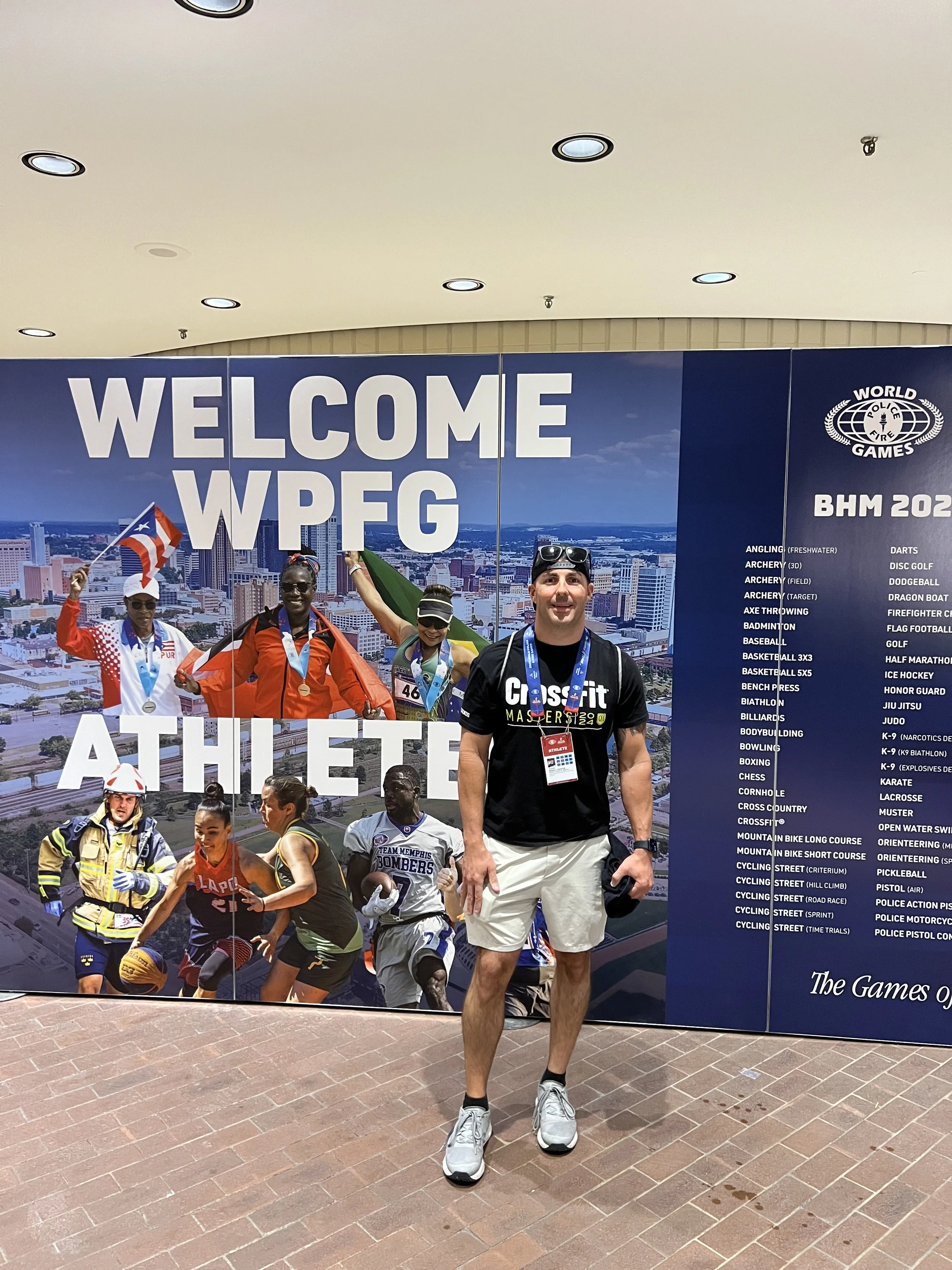 A man standing in front of a large sign at the World Police and Fire Games, with athletes in sports uniforms and medals. The sign features a cityscape and event details.