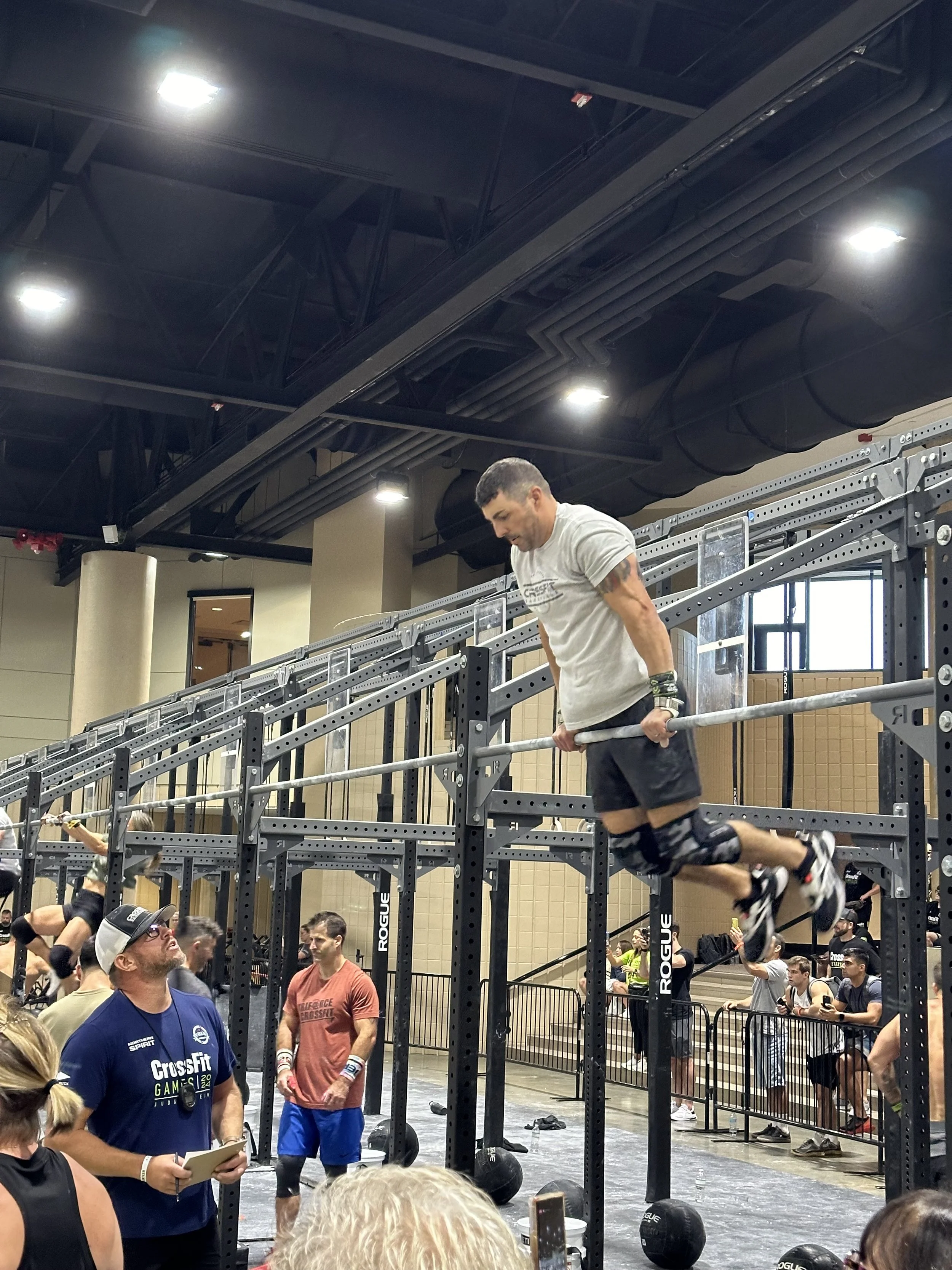 A man performing a muscle-up exercise on a pull-up bar at a CrossFit competition in an indoor gym. The gym is filled with spectators and other participants, with gym equipment and black medicine balls visible.
