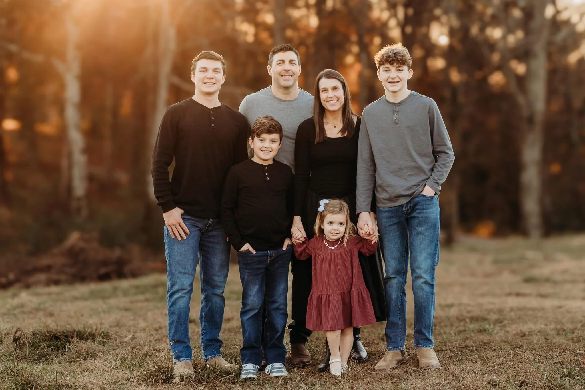 A family of six standing outdoors in a park during autumn, smiling at the camera with trees in the background.